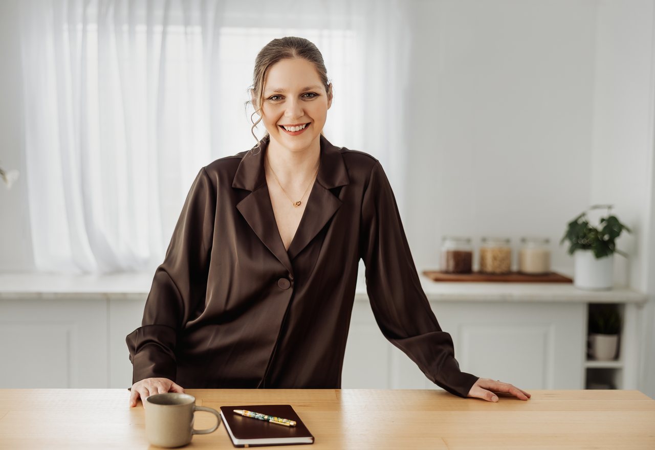 Tilly leaning on a kitchen table with a mug and notebook, smiling.