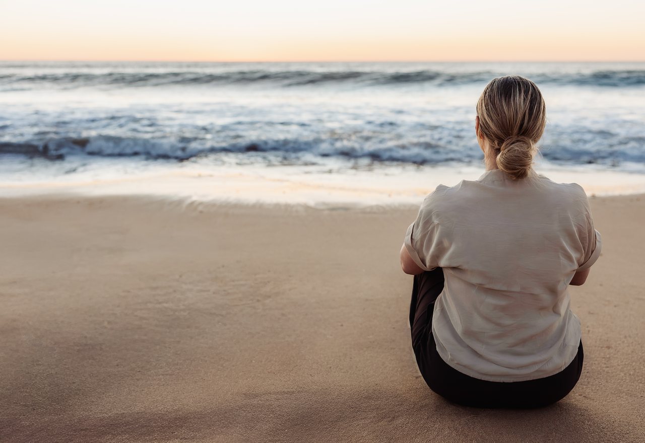 A woman sitting alone on the sand at sunrise, looking out to the ocean.