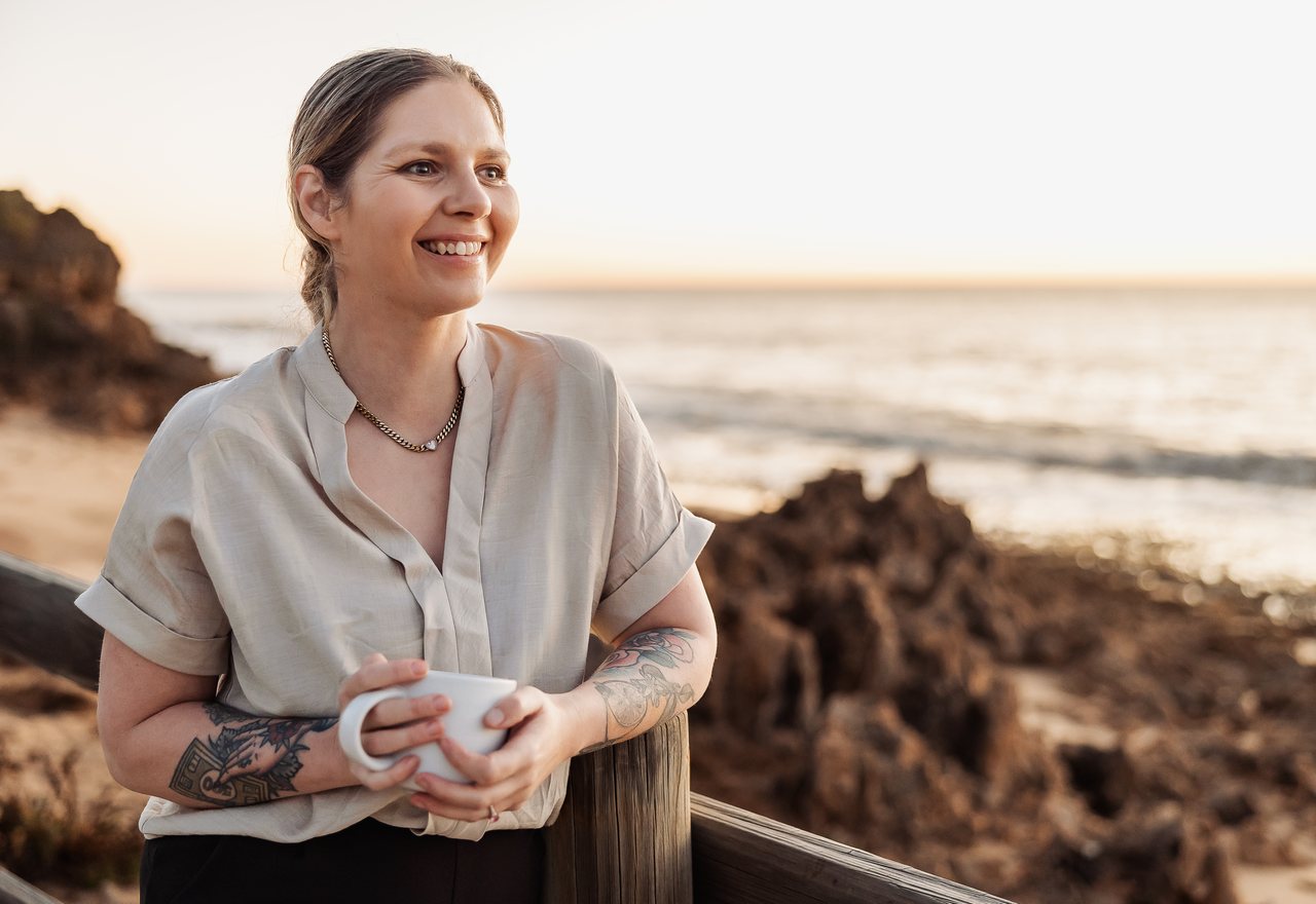 Tilly holding a mug of coffee at the coast at sunset, smiling.
